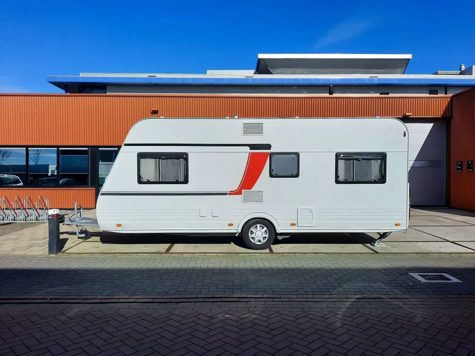 A white travel trailer with a red side flash parked alongside a workshop building, road wheel and hitch coupling in view